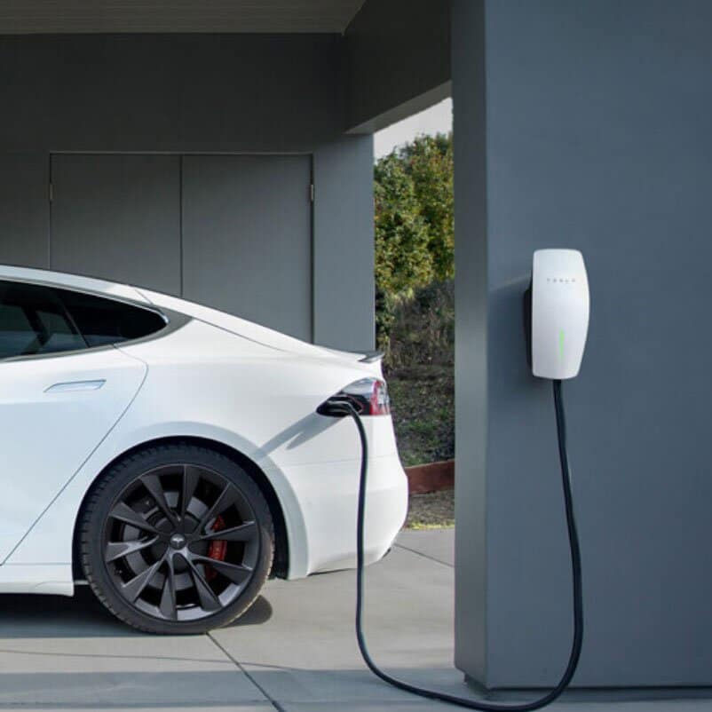 Tesla vehicle charging via a wall-mounted Tesla home charger on a private driveway, with a modern garage backdrop.